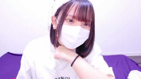 Media: A video of a young Asian woman with straight, shoulder-length dark hair, wearing a white surgical mask and white lab coat, sitting on a purple surface, indoors.