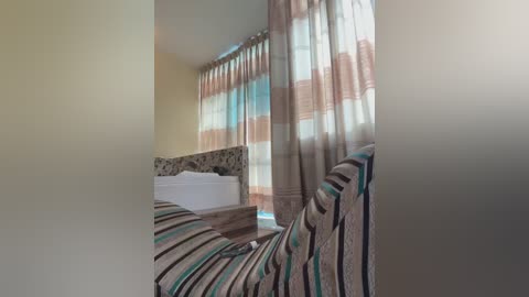 Media: Video of a cozy bedroom with beige walls, a bed with a striped coverlet, and sheer curtains with pastel stripes.