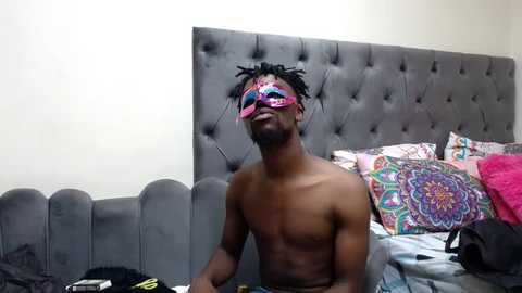 Media: Video of a shirtless, muscular Black man with dreadlocks wearing a pink and blue mask, sitting on a bed with colorful, patterned pillows and a gray, tufted headboard.