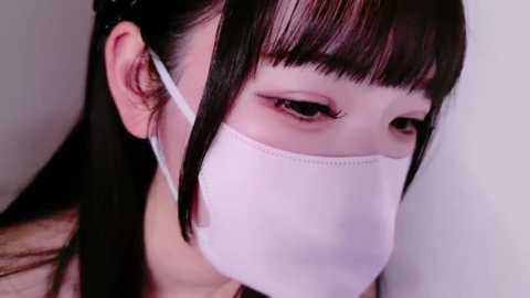 Media: Video of an East Asian woman with long black hair and bangs, wearing a white surgical mask, looking down with sad eyes, blurred background.