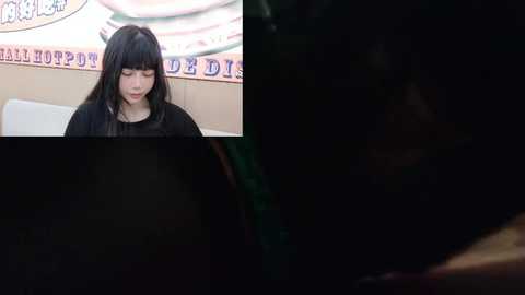 Media: A video of a young Asian woman with long black hair, wearing a black top, sitting indoors against a white wall with a colorful mural.