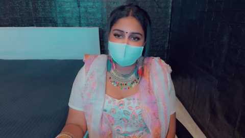 Media: Video of a South Asian woman in traditional attire, wearing a light blue face mask, floral saree, and intricate jewelry, seated on a dark floor against a brick wall.