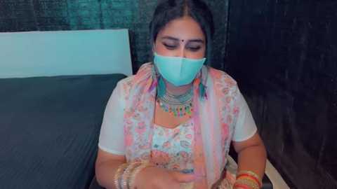 Media: Video of an Indian woman with medium skin tone, wearing a white floral sari, light blue mask, and ornate jewelry, sitting on a bed with dark sheets and a green curtain.