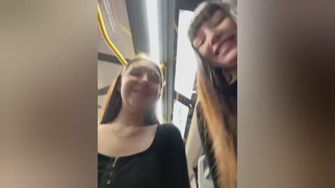 Media: Video of two smiling women in a subway car. One woman wears a black top, the other has long brown hair and a black top. Background shows yellow poles and subway seats.