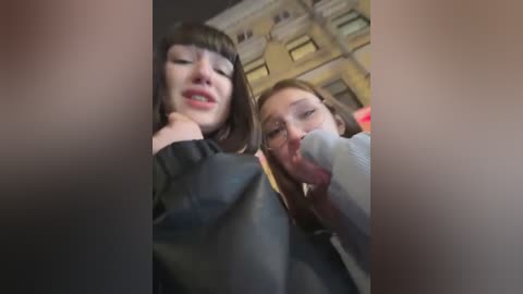 Media: Video of two young women with fair skin, one with straight black hair, the other with light brown hair. They are wearing casual clothes and appear to be kissing. Background shows a blurry, old building with a yellow facade.