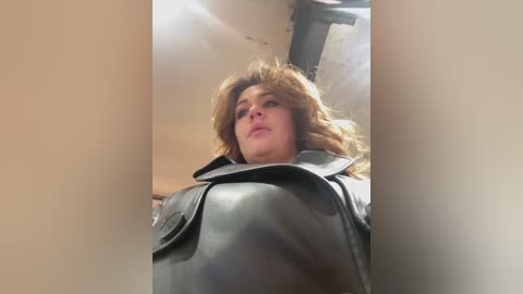 Media: Video of a woman with long, wavy brown hair, wearing a black leather jacket, looking upward with a serious expression, set against a dimly lit, industrial background with exposed pipes.