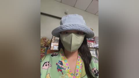 Media: Video of a person wearing a light blue bucket hat, green surgical mask, and a colorful floral-patterned shirt, standing in a store with shelves filled with various products.