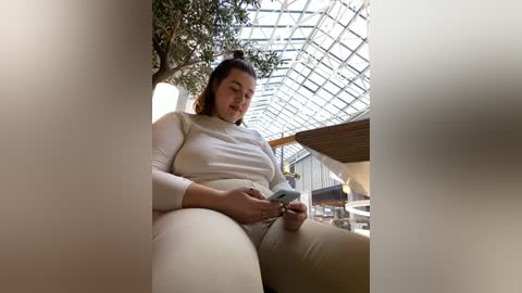 Media: A video of a plus-size woman with light skin and dark hair in a bun, wearing a white long-sleeve top and beige pants, engrossed in a phone in a sunlit, modern glass atrium.