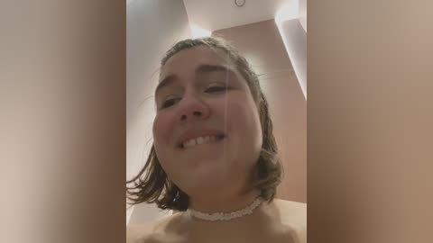 Media: Video of a young, smiling woman with light skin, shoulder-length brown hair, wearing a white choker necklace, standing in a dimly lit room with beige walls.