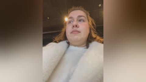 Media: Video of a young woman with light skin, long brown hair, and a white fur coat, looking upward. The background is dimly lit, with a blurred, dark ceiling.