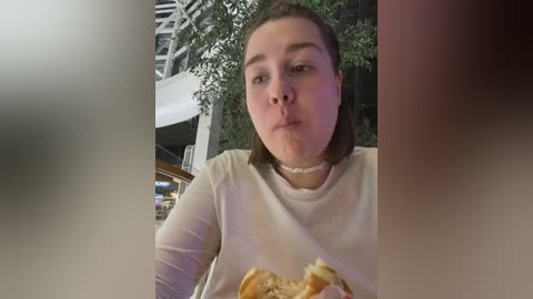 Media: Video of a plus-sized woman with short brown hair, wearing a white top, blowing a kiss to the camera, indoors with a blurred background.