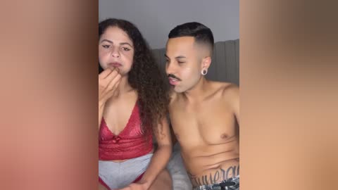 Media: Video of a light-skinned man with a mustache and earrings, shirtless, and a woman with curly hair, wearing a red lace bralette, standing close in a dimly lit room.