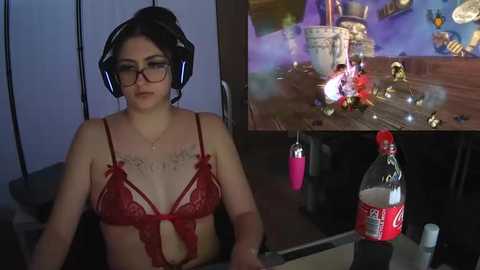 Media: Video: A woman with glasses, wearing red lace lingerie, sits in a dark room. Behind her, a video game scene shows a burning chair with a bottle of Coke.