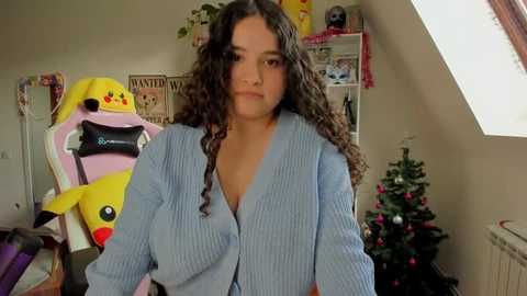 Media: Video of a young woman with curly brown hair, wearing a light blue ribbed cardigan, standing in a cluttered room with a Pikachu plush, a gaming chair, and a Christmas tree.