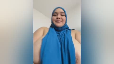 Media: A video of a smiling, plus-sized woman with a large bust wearing a blue hijab, standing in a minimalist, white-walled room.