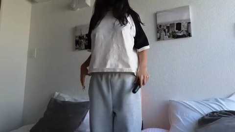 Media: Video of a woman in a white T-shirt and grey sweatpants, standing in a minimalist bedroom with white walls and two black-and-white cityscape prints.