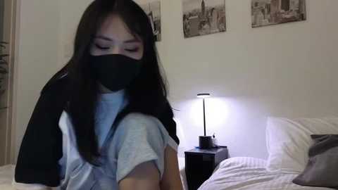 Media: Video of a young Asian woman with long black hair, wearing a face mask, light gray shirt, and black jacket, sitting on a bed in a dimly lit room with a bedside lamp and cityscape photos on the wall.