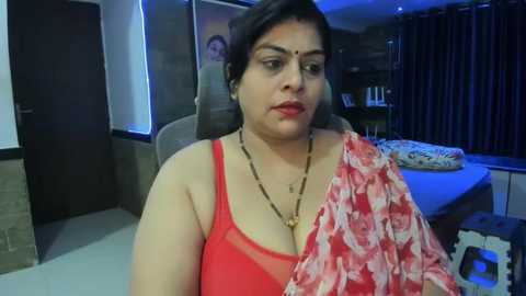 Media: Video of a plus-sized Indian woman in a red floral blouse and gold necklace, sitting on a beige chair in a dimly lit bedroom with dark curtains and a bed with white sheets.