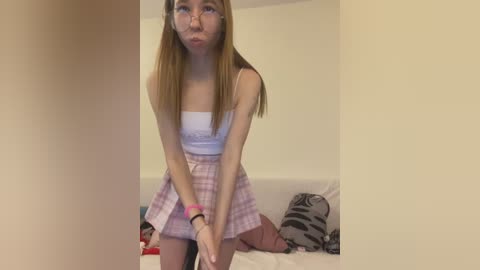 Media: Video of an Asian woman with long, straight hair, wearing a white crop top and pink plaid skirt, blowing a kiss, in a bedroom with white walls and a bed with pillows.