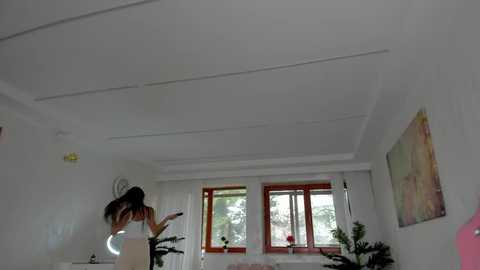 Media: Video of a modern, minimalist living room with a woman in a white top and black pants dancing energetically near a white couch. Two large windows with wooden frames and greenery outside. Artwork and potted plants adorn the walls.