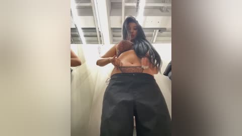 Media: Video of a young woman with long black hair, wearing a brown lace bra and high-waisted black pants, standing in a narrow, dimly lit corridor with white walls and exposed ceiling.