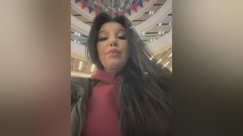 Media: Video of a young woman with long black hair, wearing a red turtleneck sweater and a gray jacket, standing in a modern, circular building with a glass ceiling.