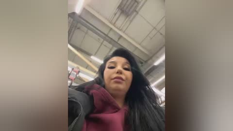 Media: Video of a young woman with long black hair and light skin, wearing a maroon hoodie, standing in an industrial setting with exposed ceilings and fluorescent lights.