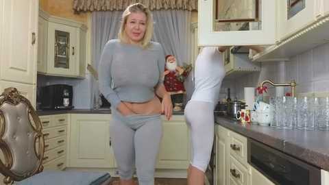 Media: Video of a blonde woman in light blue workout gear adjusting her pants in a cozy, cream-colored kitchen with floral curtains and a decorative chair.