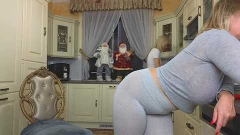 Media: Video of a modern kitchen with a large woman in light blue pajamas bending over a counter, Santa Claus figurines on shelves, and a blonde woman in the background.