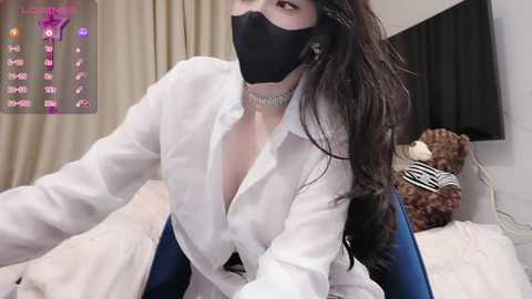 Media: A video of a woman with long black hair, wearing a white shirt, black mask, and a choker, sitting on a bed with teddy bears, in a dimly lit bedroom.