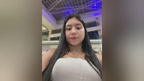 Media: Video of a young Latina woman with long, straight black hair, wearing a white tube top, standing indoors with a modern, high ceiling, and visible metal beams in the background.