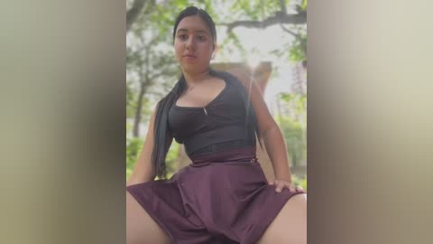 Media: Video of a Latina woman with long black hair, medium skin tone, wearing a dark gray tank top and maroon skirt, seated outdoors in a garden with green foliage and blurred background.