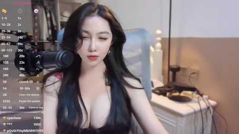 Media: Video of a young Asian woman with long black hair, wearing a revealing black top, sitting in a dimly lit room with a white desk and a laptop, capturing a live stream on a smartphone.