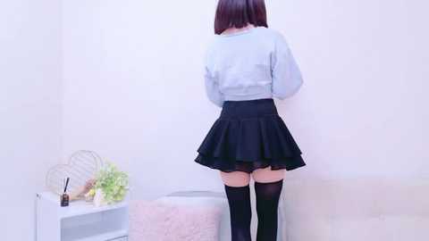 Media: Video of a woman with straight, shoulder-length brown hair, wearing a light blue blouse, black pleated skirt, and thigh-high black stockings, standing against a white wall in a minimalist room with a white chair and a shelf holding a vase and a bottle.