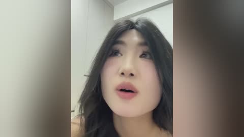 Media: Video of an Asian woman with long, black hair, fair skin, and full lips, looking surprised and slightly open-mouthed. The background is a plain, white wall with a blurred doorway, suggesting an indoor setting.