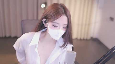 Media: Video of an Asian woman in a white lab coat, white face mask, and headphones, intently reading a tablet. Background features beige curtains and a gray chair.