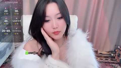 Media: Video of an East Asian woman with long black hair, wearing a white fur coat, resting her head on her hand in a cozy room with pink curtains and a patterned rug.