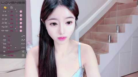 Media: A video of a young Asian woman with long black hair, wearing a light blue bra, standing in a modern, bright living room with wooden stairs and white walls.