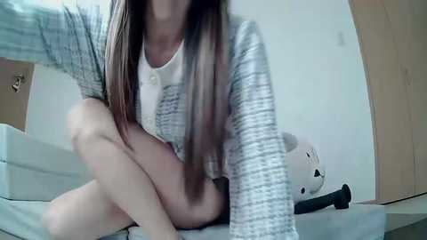 Media: Video of a young woman with long, straight brown hair, wearing a gray tweed jacket and white top, sitting on a bed with a stuffed animal.