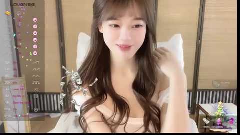 A video of an Asian woman with long brown hair, fair skin, and a soft smile, sitting on a bed with a white comforter, in a modern room with wooden blinds.