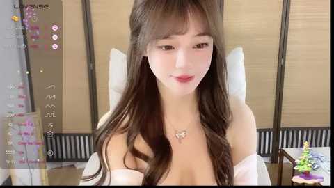 Video of a young Asian woman with long brown hair, wearing a white off-shoulder top, sitting on a white chair in a modern bedroom with wooden sliding doors.