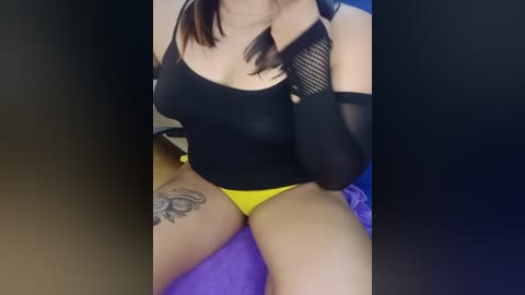 Media: Video of a woman with fair skin and brown hair, wearing a black mesh sleeve, black top, and yellow panties, sitting on a purple surface, showcasing a thigh tattoo.