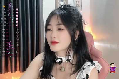 Media: A video of a young East Asian woman with pale skin and long black hair, wearing a white and black polka dot dress with a choker and a bell pendant. She sits in a pink chair, looking serene. Background includes black curtains and a blurry room.