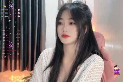 Media: A video of an East Asian woman with long black hair and fair skin, wearing a white knit cardigan, sitting indoors with a pink chair and dark curtains.