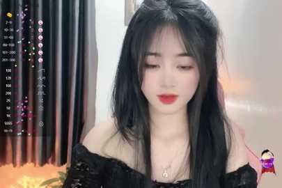 Media: Video of a fair-skinned East Asian woman with long black hair, red lipstick, and a black off-shoulder top, indoors with a soft-focus background.