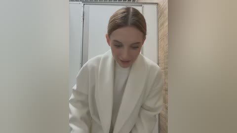 Media: Video of a young woman with light skin and straight, light brown hair, wearing a white bathrobe, leaning forward in a beige-tiled shower stall with a white shower curtain.