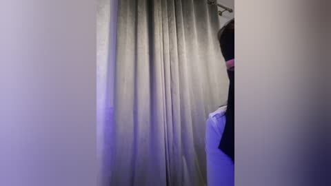 Media: Video of a person standing in a dimly lit room with a gray curtain, partially visible in the background. The person's face is partially obscured, with a hint of a pink scarf around their neck.