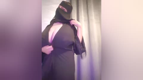 Media: Video of a woman with light skin, wearing a black niqab and flowing black dress, adjusting her veil in a dimly lit room with gray curtains.