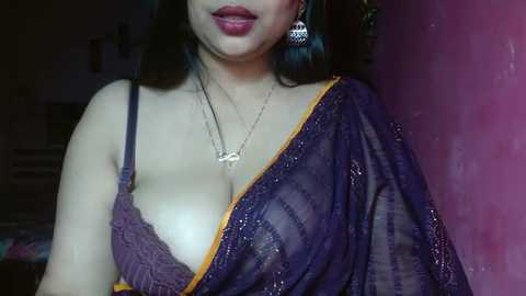 Media: A video of a woman with fair skin, wearing a purple saree with a yellow border, and a lace bra, standing against a pink wall. She has long black hair and wears dangling earrings.