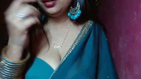 Media: Video of a South Asian woman with medium skin tone, wearing a teal saree with golden embroidery, turquoise earrings, and a silver necklace. She has a confident expression, talking on a phone, with a brown bangle on her wrist.